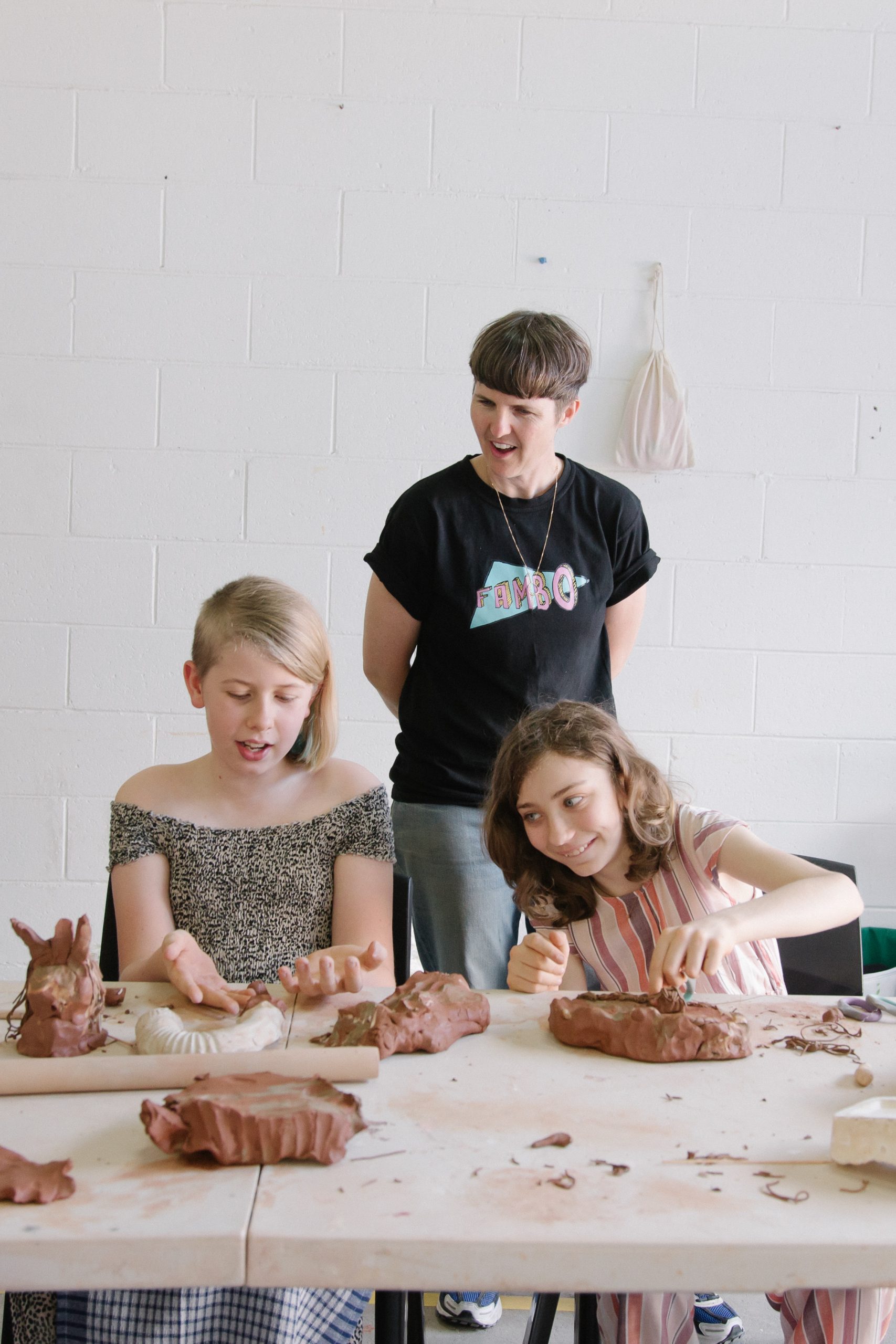Two kids are creating clay figures, with an adult looking impressed behind them, wearing a Fambo tshirt